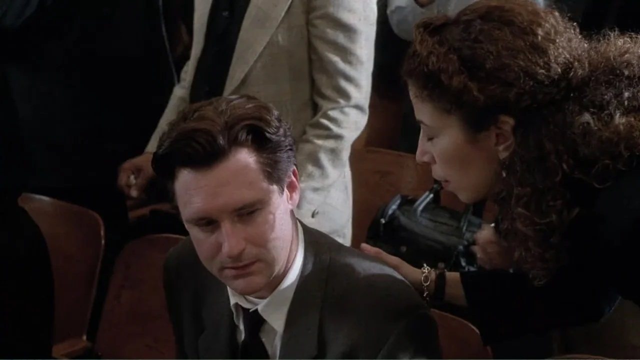 Mistrial (1996) movie still: medium — A crowded auditorium contains rows of dark wooden folding seats occupied by seated people. A woman leans over…