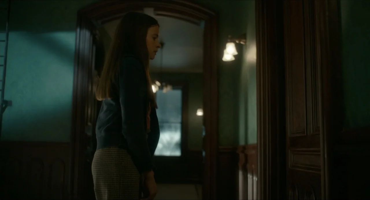 IF (2024) movie still: medium — A dimly lit indoor hallway with dark wood paneling and green wallpapered walls. A young girl in a hoodie stands in…