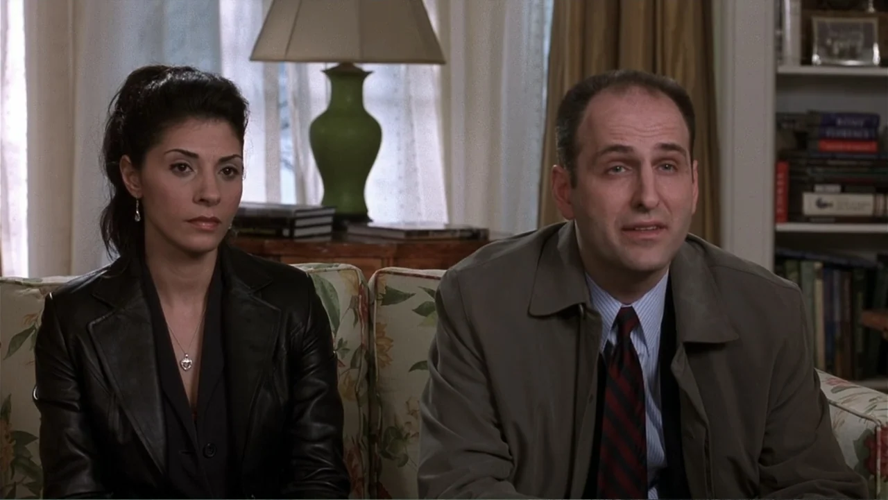 Analyze That (2002) movie still: medium — A well-lit residential room features a floral sofa positioned in front of a window. A man in a trench coat…