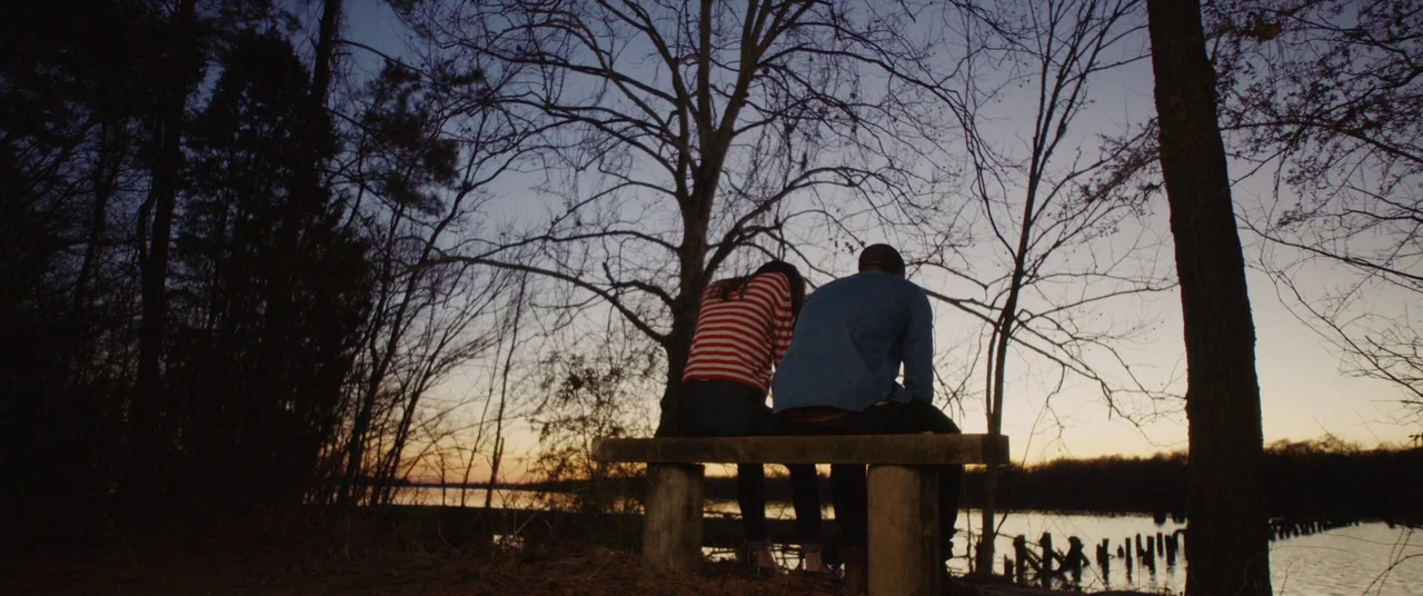 Get Out (2017) movie still: wide — A wooden bench sits on a grassy bank overlooking a calm river at dusk. Two people sit together on the bench with…