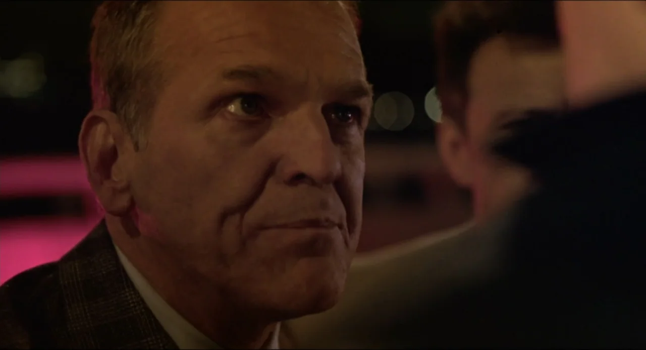 Cop Land (1997) movie still: closeup — A dimly lit bar setting with blurred lights visible in the background. An older man sits while observing…