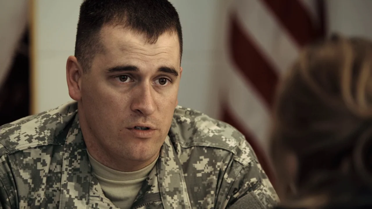 Winter’s Bone (2010) movie still: closeup — An office interior features a blurred American flag visible in the background. A soldier in uniform…