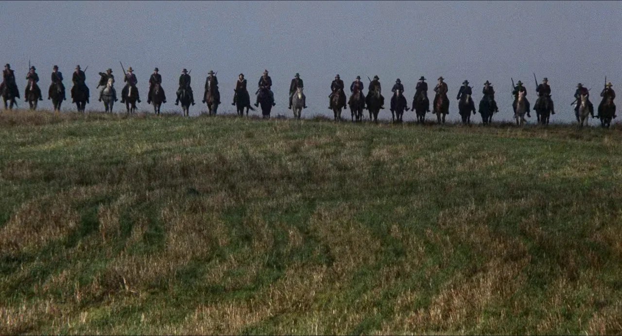 Breaker Morant (1980) movie still: wide — A grassy hill rises against a overcast sky in an open landscape. Numerous men on horseback sit in a line…