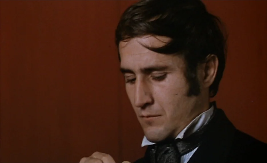 The Immortal Story (1968) movie still: closeup — A man stands before a flat reddish wall in a dimly lit interior. He keeps his gaze lowered while…