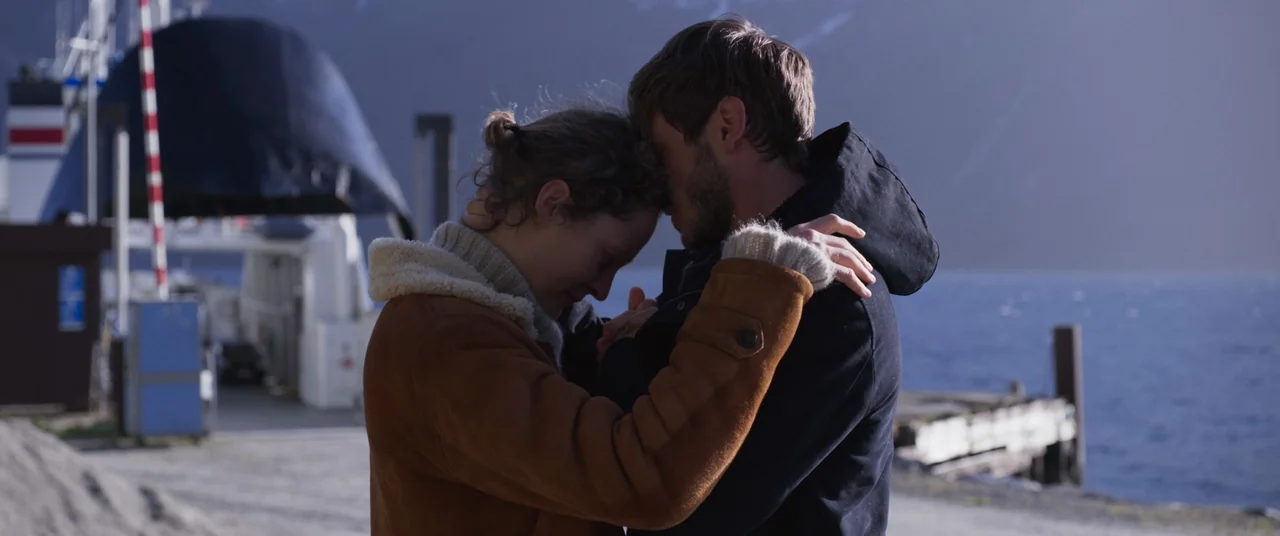More Than Ever (2022) movie still: medium — A ferry terminal with a large ship docked at a wooden pier by water. Two people stand in the foreground…
