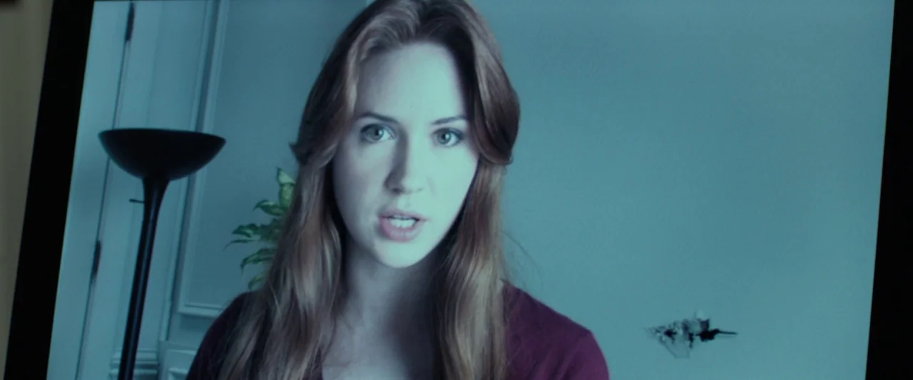 Oculus (2013) movie still: closeup — A digital screen displays a video feed of a woman in a dimly lit room. She looks toward the camera while…