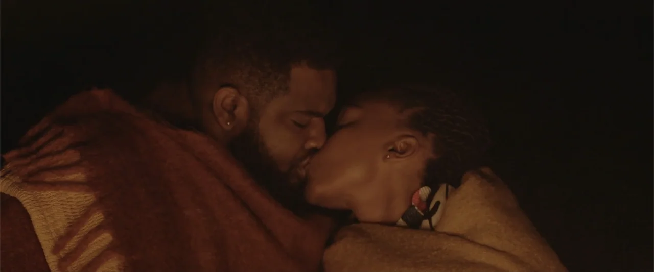 A Scam Called Love (2025) movie still: closeup — A dark, dimly lit interior space with the two subjects under heavy blankets. A man and a woman are…