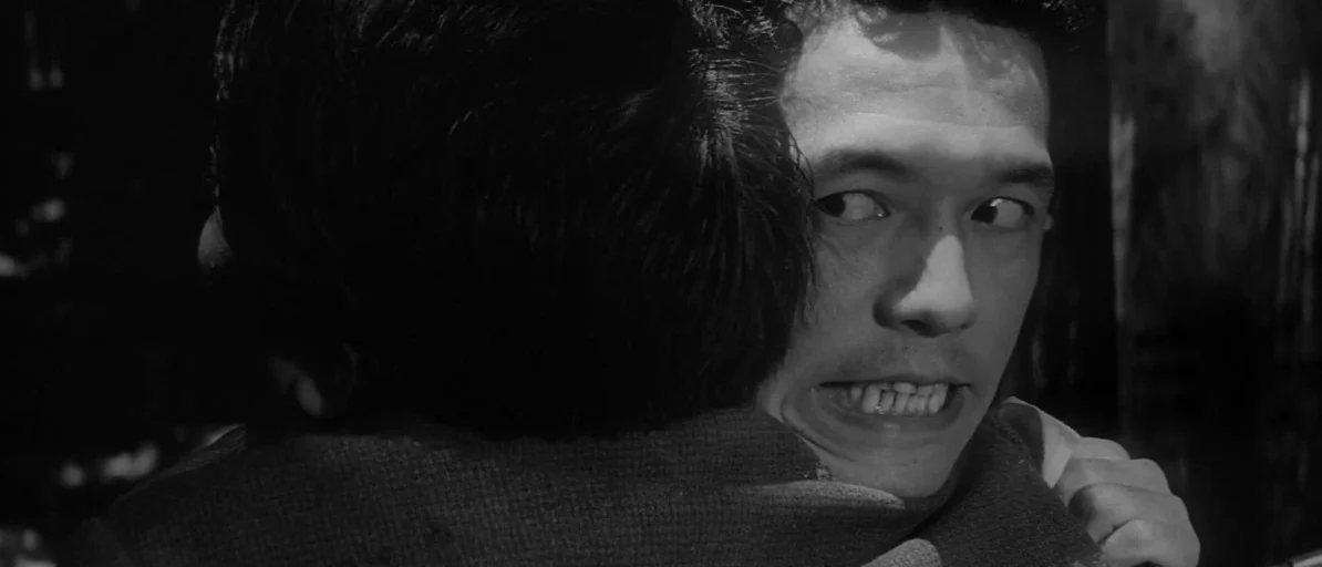 Branded To Kill (1967) movie still: closeup — A dark, dimly lit environment where two figures are locked in a close embrace. The face of one man is…
