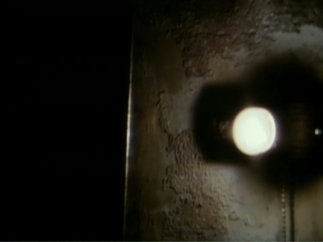 Curtains (1995) movie still: closeup — A close-up shot of a rough textured wall surface in a darkened interior. A single bright practical bulb…