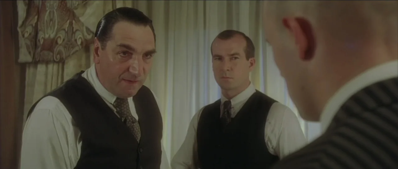 Legionario (2025) movie still: medium — An interior office space with draped windows and classic 1930s business attire. Two men stand in…