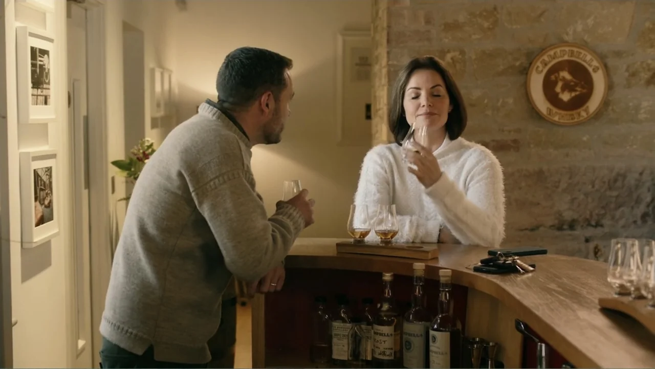 A Scottish Love Scheme (2024) movie still: medium — A well-lit interior features a curved wooden bar counter with stone walls behind it. A man and a…