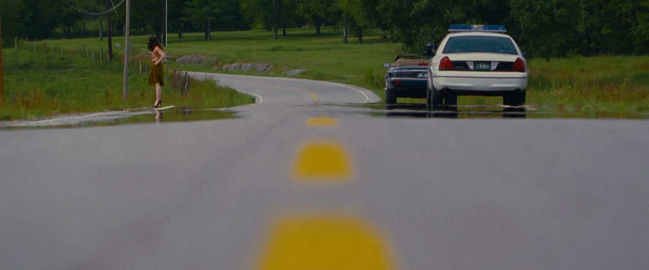 Stoker (2013) movie still: wide — A rural paved road winds through green fields with trees in the background. A lone woman stands on the side of the…