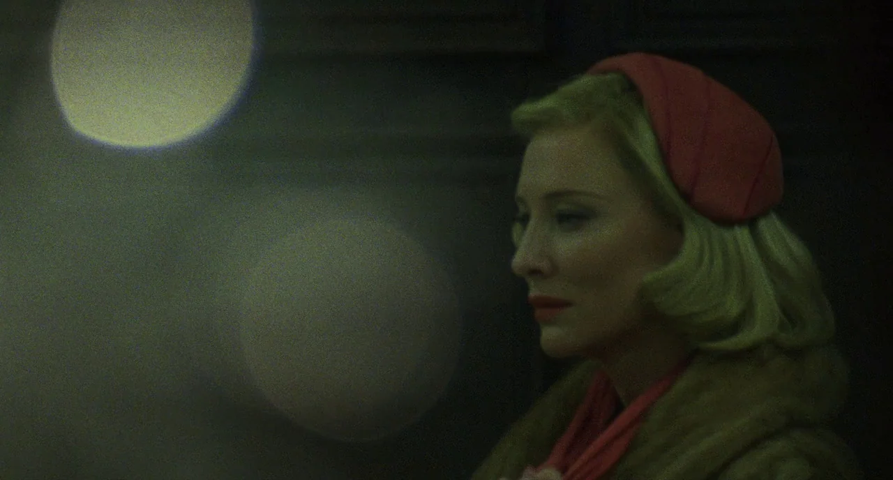 Carol (2015) movie still: closeup — A night city street is obscured by dark shadows and large blurry out-of-focus highlights. A woman stands in…