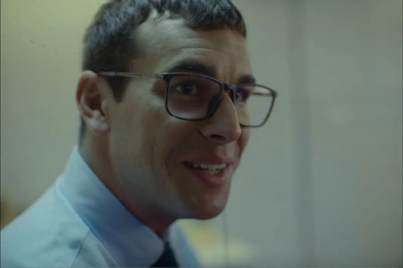 Cross the Line (2020) movie still: closeup — An interior office space with plain, light-colored walls in the background. A man wearing glasses and a…