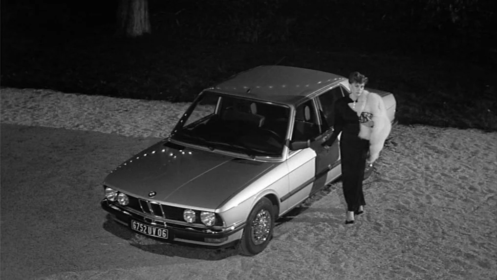 Under The Cherry Moon (1986) movie still: wide — A dark, unpaved outdoor surface at night serves as the backdrop for a silver sedan. A woman in a…