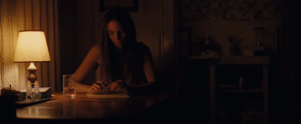 Ain’t Them Bodies Saints (2013) movie still: medium — A dimly lit room features a table lamp casting a warm glow on writing materials. A woman sits…