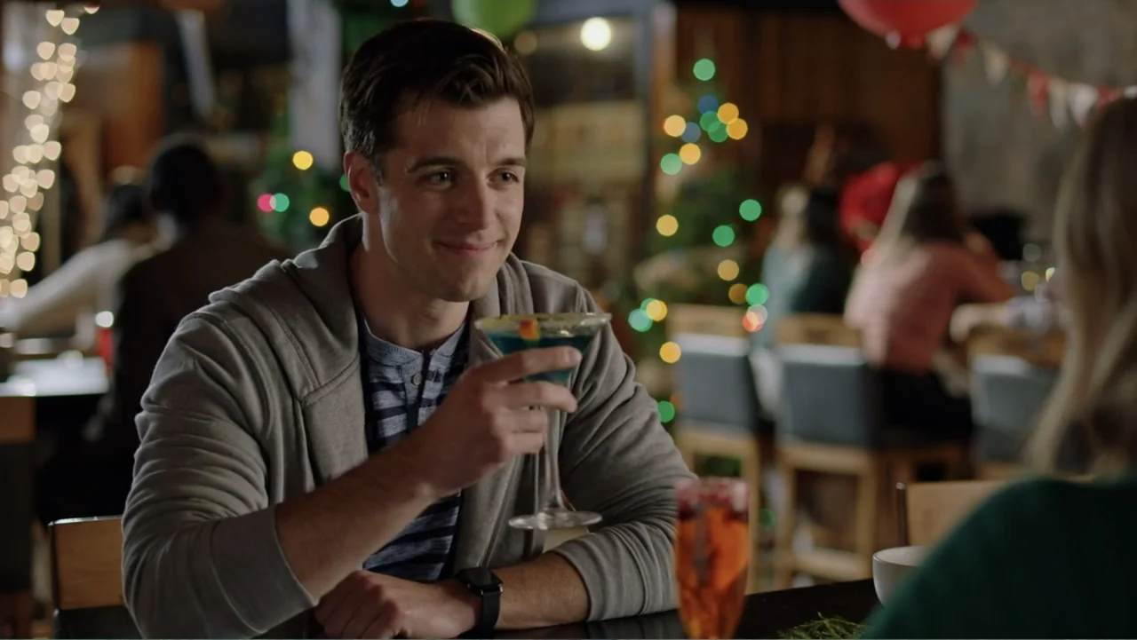 Ghosts of Christmas Past (2021) movie still: medium — A festive bar setting with Christmas lights glowing in the blurred background. A man in a grey…