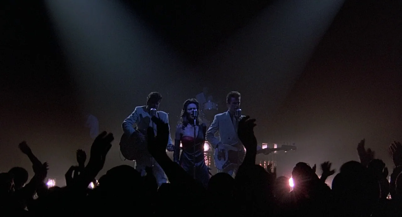 Streets of Fire (1984) movie still: wide — A dark concert hall stage is illuminated by focused overhead lights during a performance. A band stands…