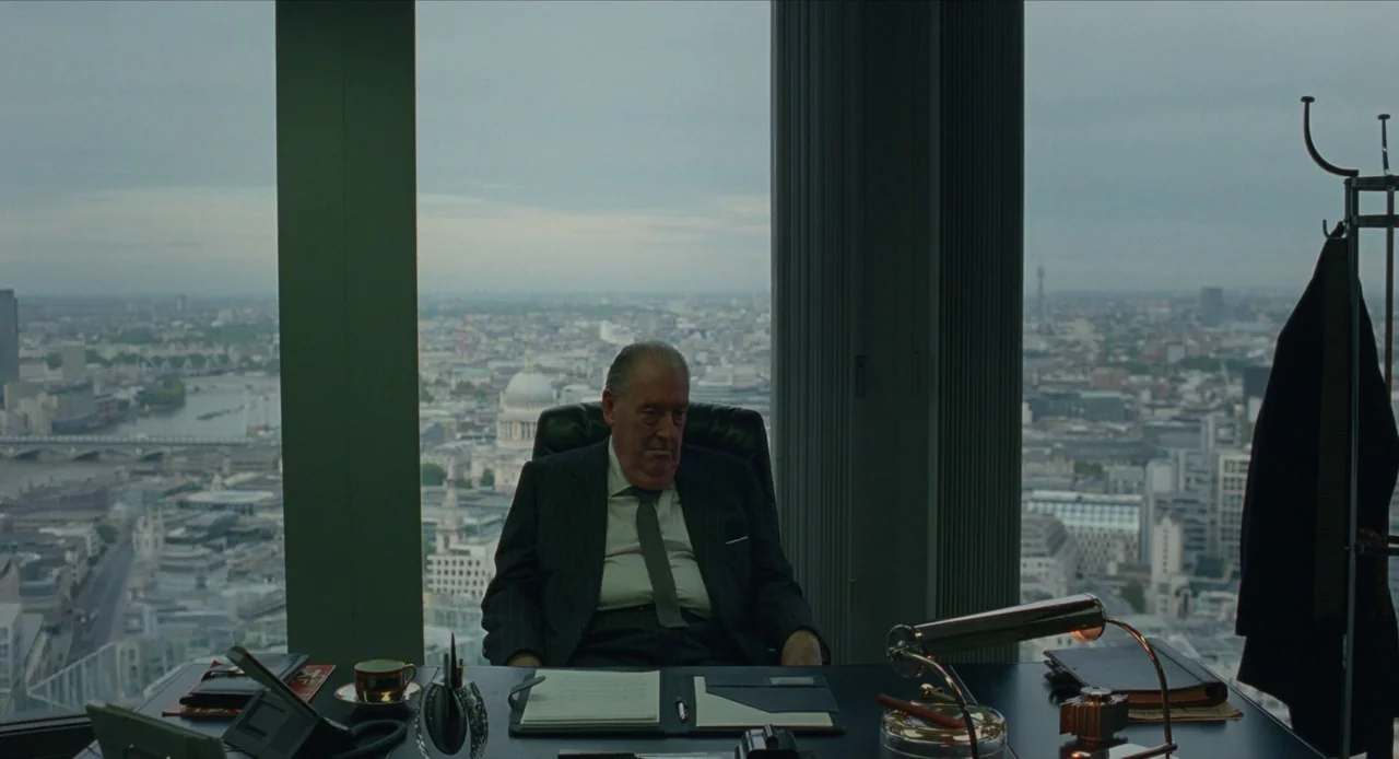 The Nest (2020) movie still: medium — A high-rise executive office with a panoramic view of London featuring St Paul's Cathedral. A man sits behind…