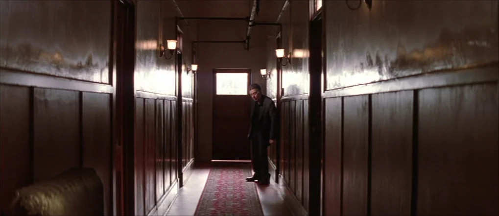 Insomnia (2002) movie still: wide — A long, dim hallway with dark wooden wall paneling and a red patterned runner carpet. A lone man in a dark suit…