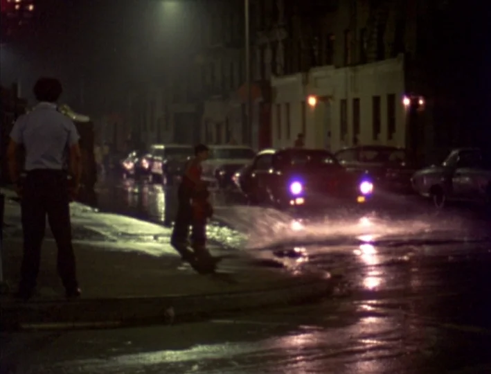 News From Home (1977) movie still: fullbody — A rain-slicked city street at night features buildings under dim street lighting. A man stands on the…