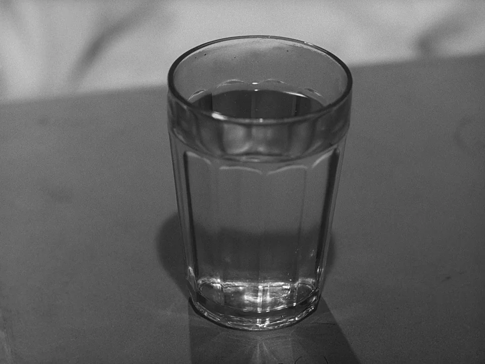 Brink of Life (1958) movie still: closeup — A faceted glass filled with liquid sits on a flat, smooth surface. The scene is illuminated with clear…