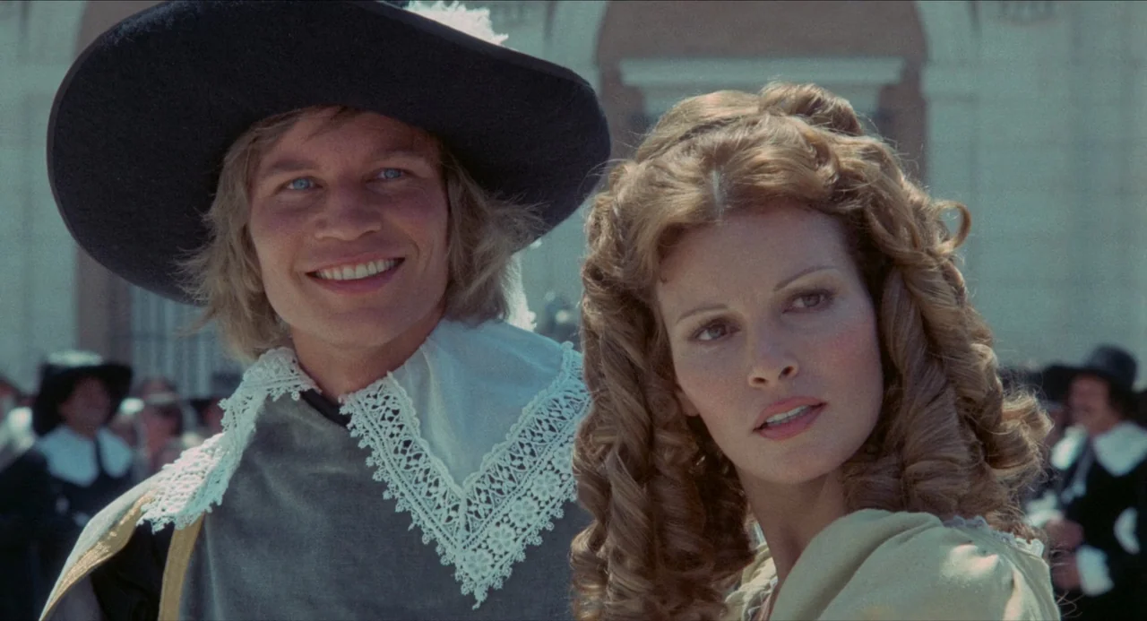 The Three Musketeers (1973) movie still: medium — A public outdoor plaza with out-of-focus background figures in historical period attire. A man in…