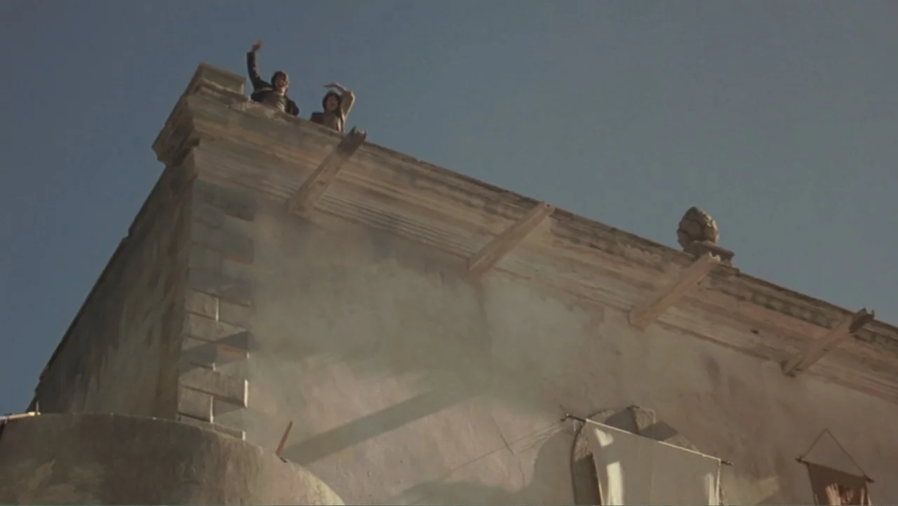 The Mask of Zorro (1998) movie still: wide — A low-angle view of a beige stone building with wooden roof supports. Two people stand on the edge of…