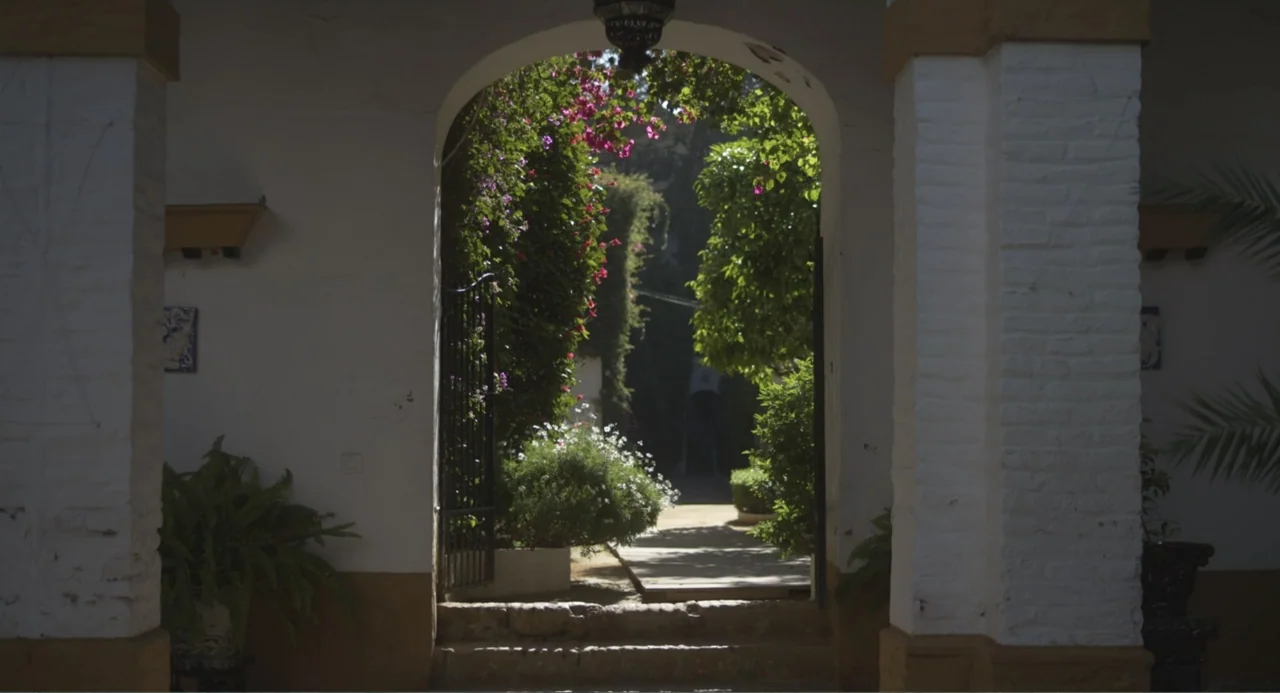 Los días azules (2025) movie still: establishing — A white stucco archway leads into a sunlit courtyard filled with lush green vegetation. Potted…