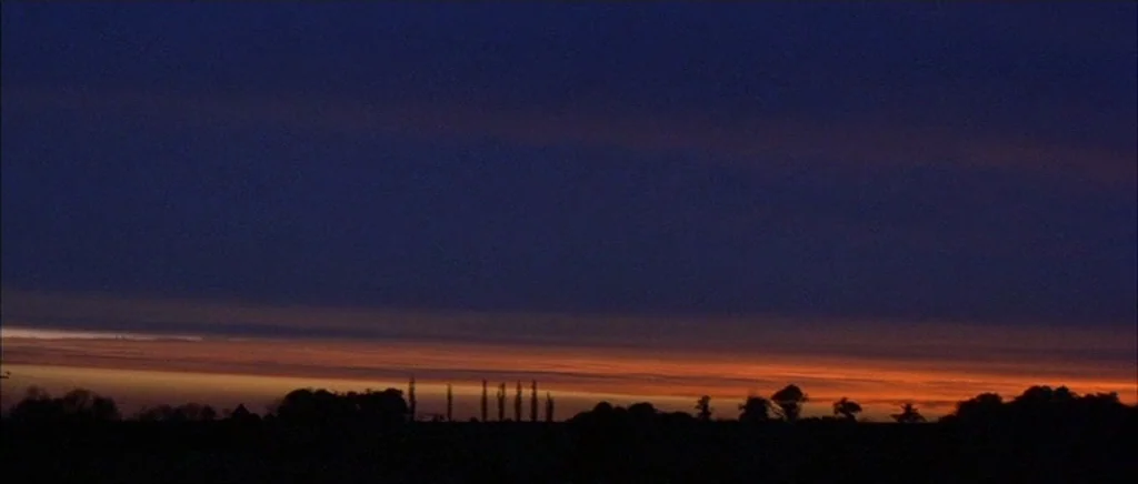 Jude (1996) movie still: establishing — A dark, silhouetted treeline sits beneath a vast twilight sky. A sliver of warm orange light stretches…