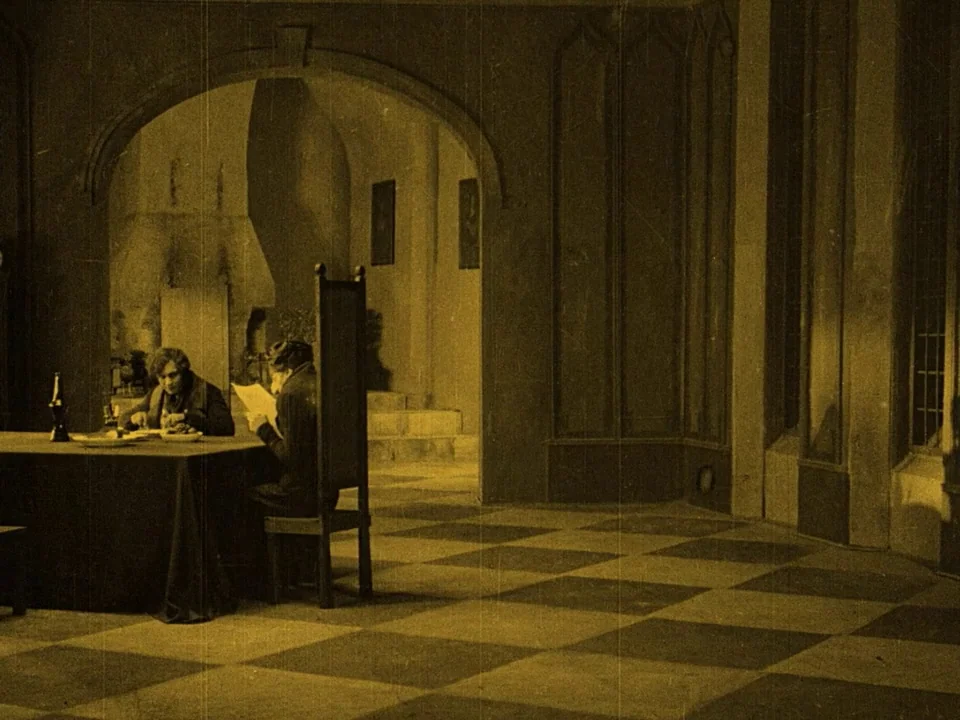 Nosferatu (1922) movie still: wide — A spacious dining hall features a high-backed chair, a checkered floor, and stone arches. Two men sit at a long…