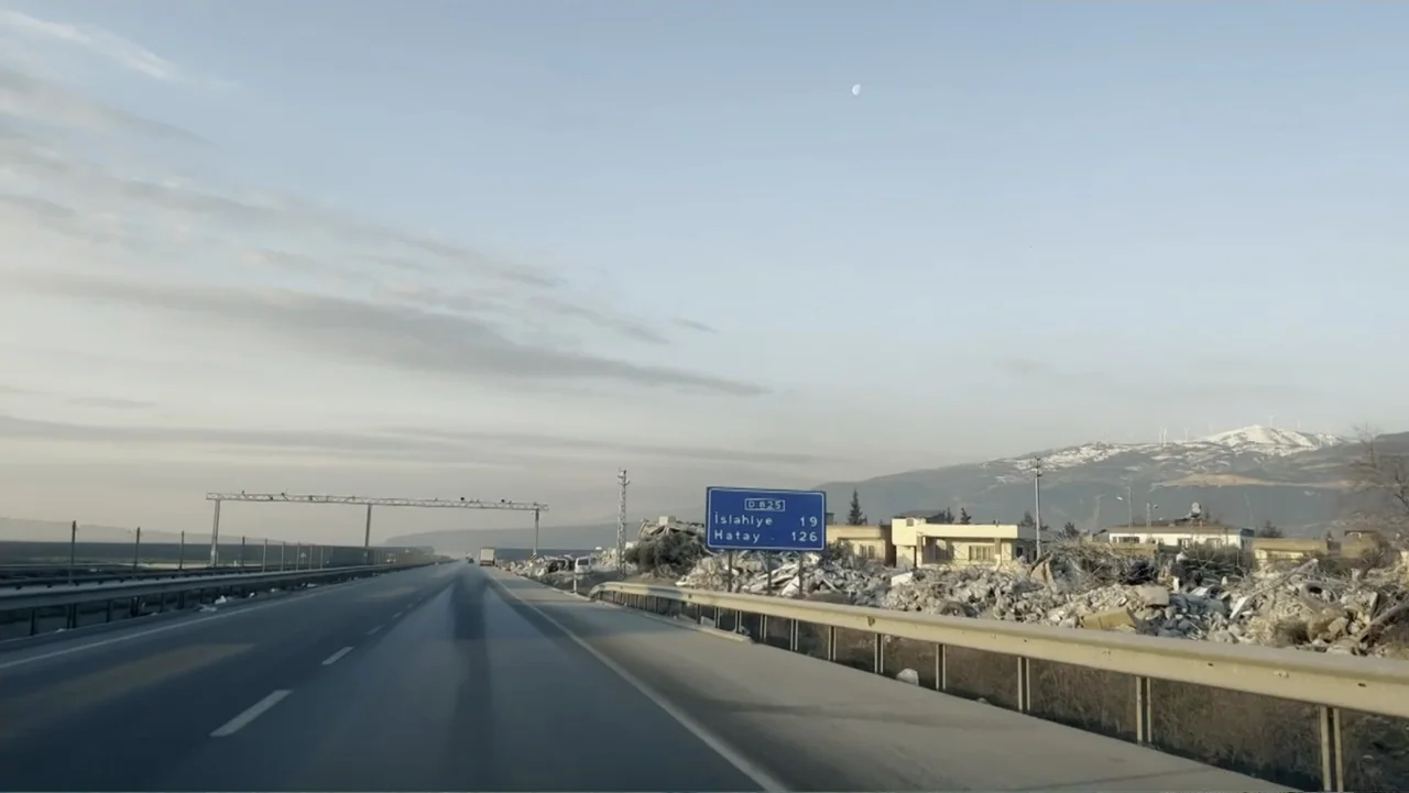 Death Without Mercy (2024) movie still: establishing — A wide paved highway stretches forward past piles of building debris and rubble. Mountains…