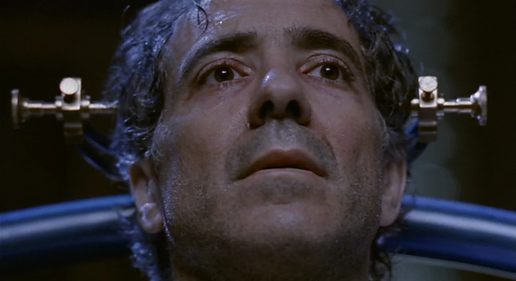 Kafka (1991) movie still: closeup — A dimly lit room holds a man secured within a metallic head restraint device. The man gazes upward with a…