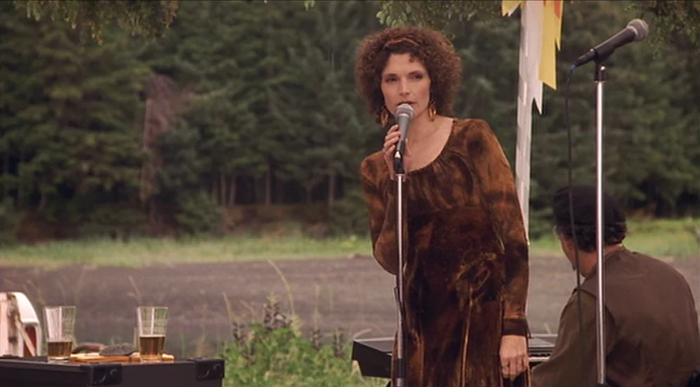 Limbo (1999) movie still: medium — A woman stands on an outdoor stage in front of a dense pine forest. A man sits nearby behind a keyboard as she…