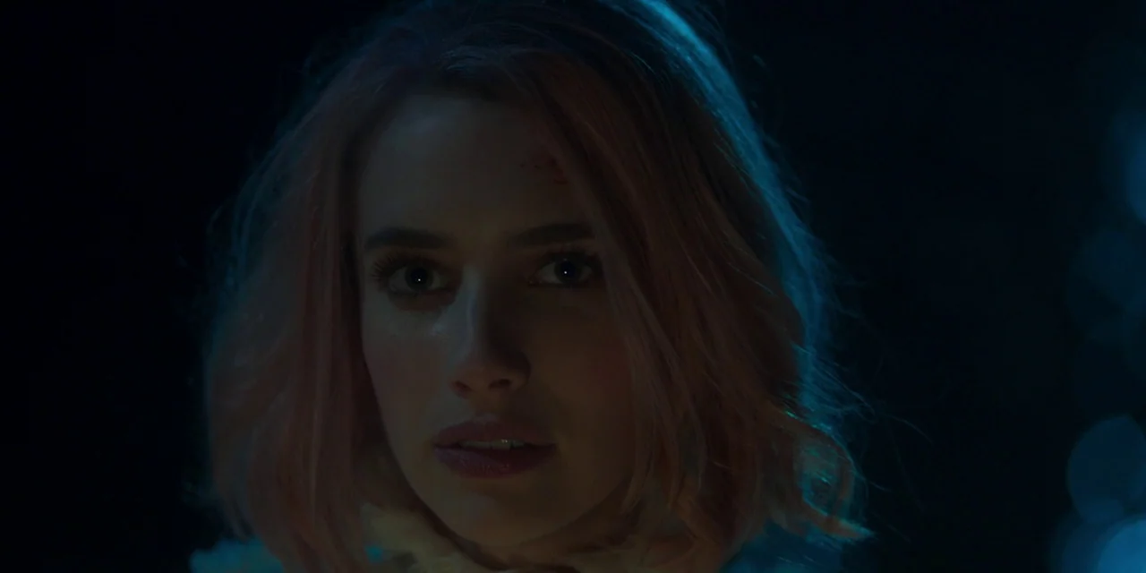 Paradise Hills (2019) movie still: closeup — A dark, undefined environment surrounds a young woman with pink hair. She gazes ahead with a neutral…