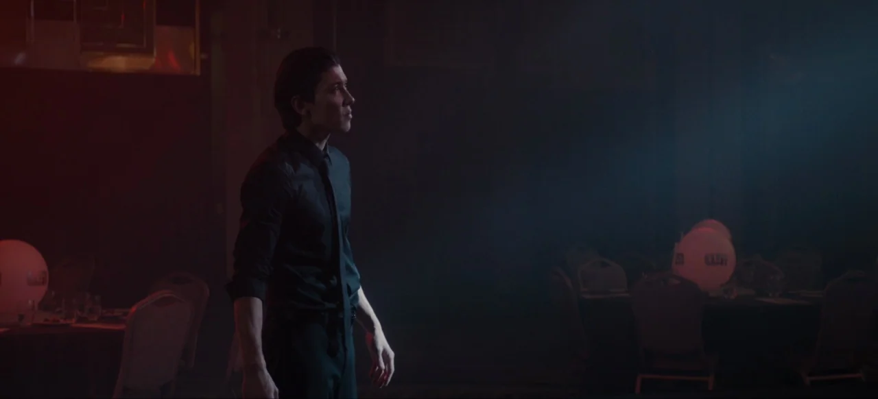 Housewife (2017) movie still: medium — A dark event space contains round tables with white spheres and dim lighting. A man stands alone in the…