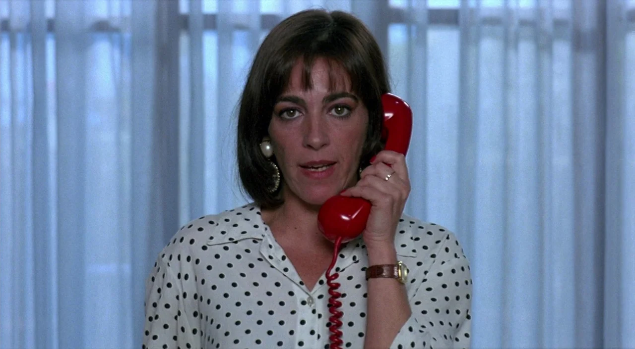 Women on the Verge of a Nervous Breakdown (1988) movie still: closeup — A woman stands before a white curtained backdrop in an indoor setting. She…
