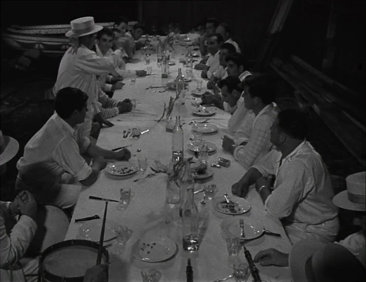 La Pointe Courte (1955) movie still: wide — A long banquet table is set for a dinner with many men dressed in light-colored clothing. The men sit…