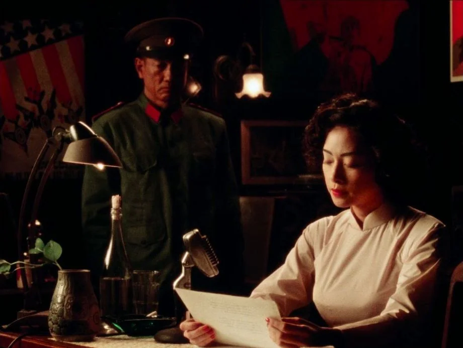 Da 5 Bloods (2020) movie still: medium — A dimly lit office features vintage posters on the wall and a desk microphone. A woman in a traditional…