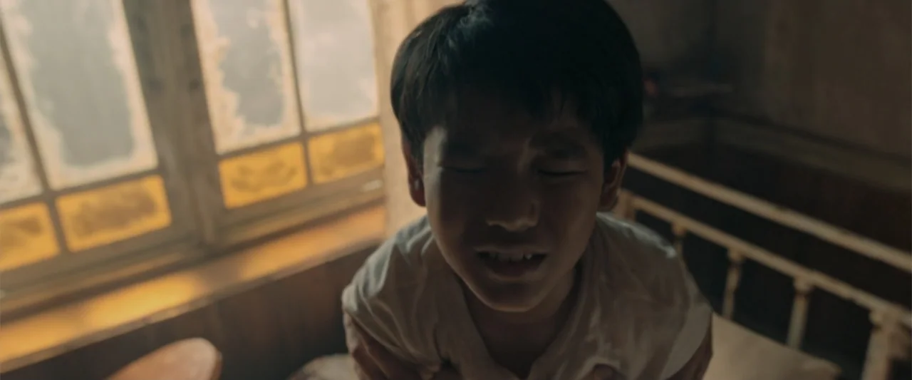 The Unholy (2022) movie still: closeup — A dimly lit bedroom with aged walls and a window with yellow-tinted glass panes. A young east asian boy in…