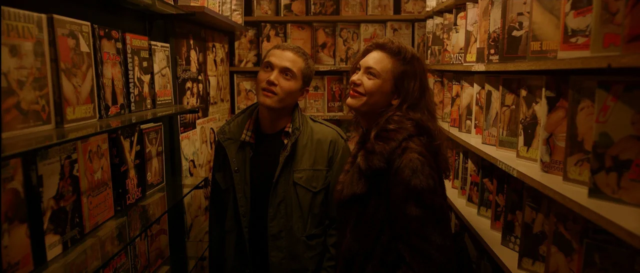 Love (2015) movie still: medium — A narrow aisle of a vintage adult video store features packed shelves of media. A young couple walks through the…