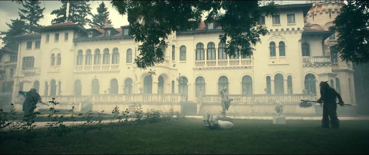 The Toxic Avenger (2023) movie still: wide — A sprawling stone mansion features multiple arched windows and a white balustrade terrace. Two costumed…