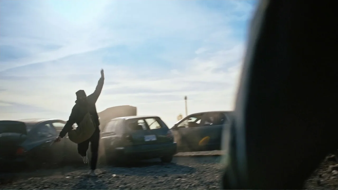 Bullet Proof (2022) movie still: wide — A dusty outdoor lot is filled with abandoned vehicles under a bright sky. A man in black runs through the…