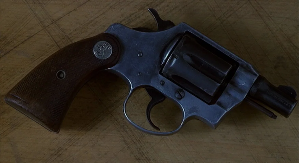 Misery (1990) movie still: closeup — A dark revolver with a wooden grip rests flat on a scarred wooden table. Deep scratches cover the light brown…