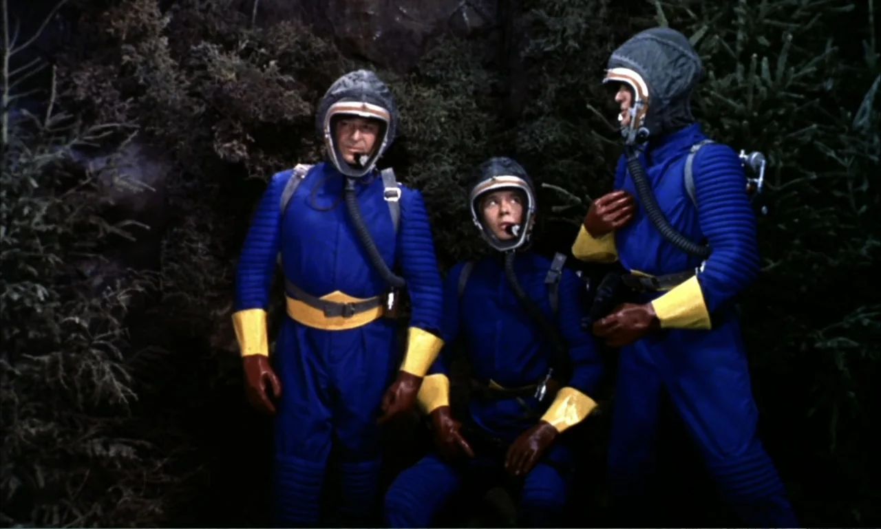 Journey to the Seventh Planet (1962) movie still: medium — A dense patch of dark evergreen trees fills the background behind three space explorers.…