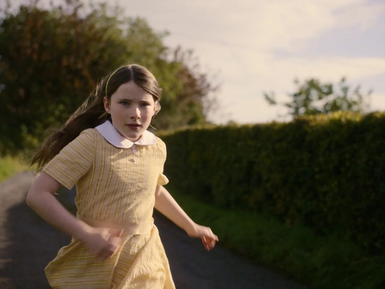 The Quiet Girl (2022) movie still: medium — A narrow asphalt lane is lined by a thick, manicured green hedge under daylight. A young girl in a…