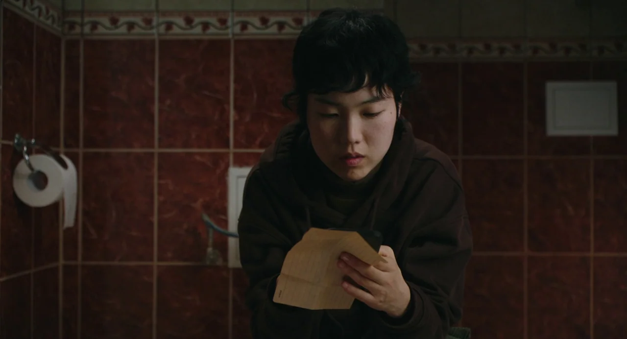 Return To Seoul (2022) movie still: medium — A tiled room features deep red walls with a decorative floral border near ceiling. A person sits while…
