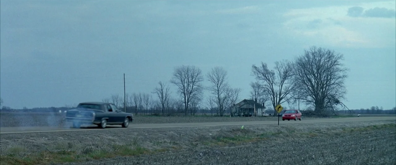 Ballast (2008) movie still: wide — A flat rural landscape features a paved road running through barren fields and trees. A dark sedan drives away…