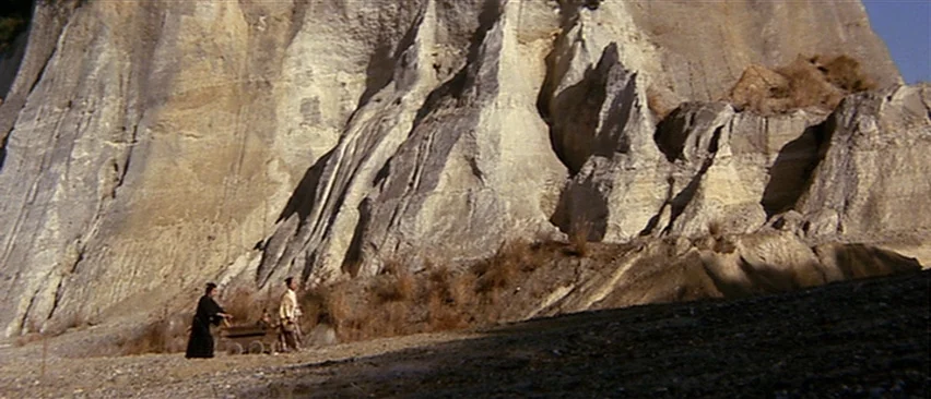 Lone Wolf and Cub: Baby Cart in Peril (1972) movie still: wide — A vast, eroded pale rock cliff towers over a dry, pebble-strewn riverbed. Two men…