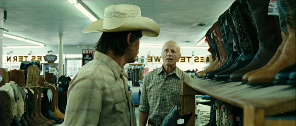 No Country For Old Men movie still: medium — A retail shop interior contains long shelves lined with leather western-style boots. A man wearing a…